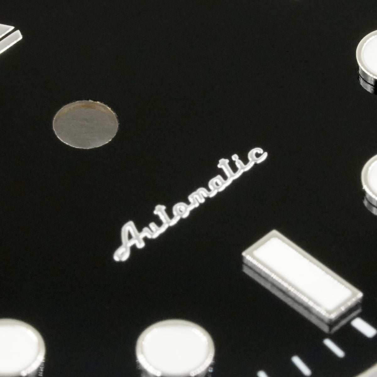 Macro of the applied polished-silver Automatic script on the glossy black enamel Submariner II dial, beside a lumed baton marker and the dial's centre hole