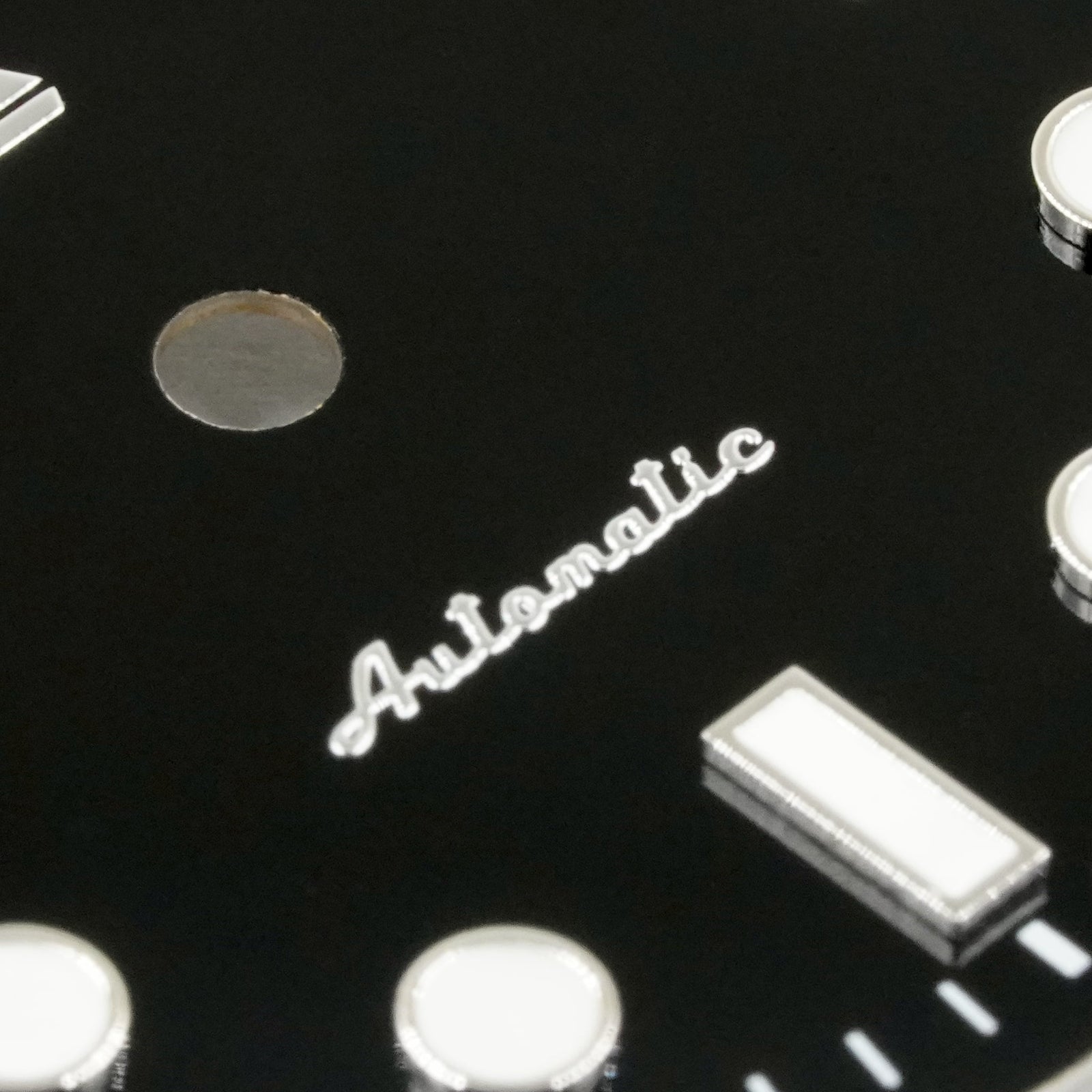 Macro of the applied polished-silver Automatic script on the glossy black enamel Submariner II dial, beside a lumed baton marker and the dial's centre hole